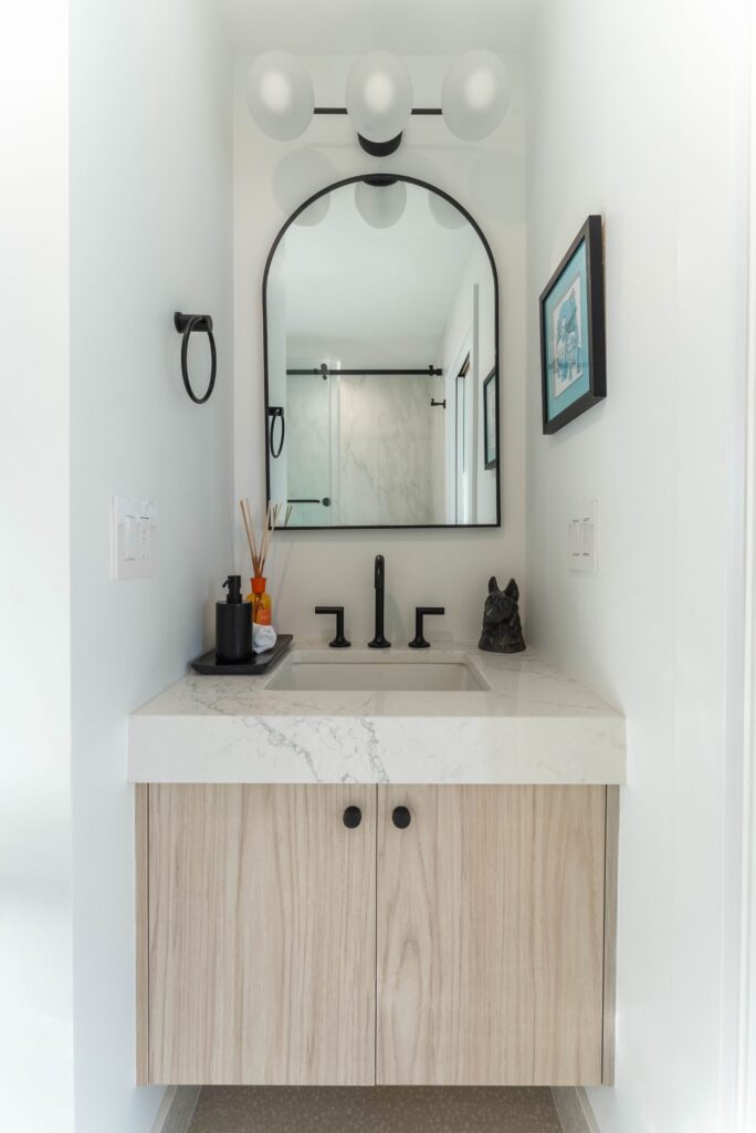 A modern bathroom vanity with a light wood cabinet, white marble countertop, black fixtures, a large arched mirror, and wall-mounted lights. A framed picture and a glass shower with black hardware are visible in the background.