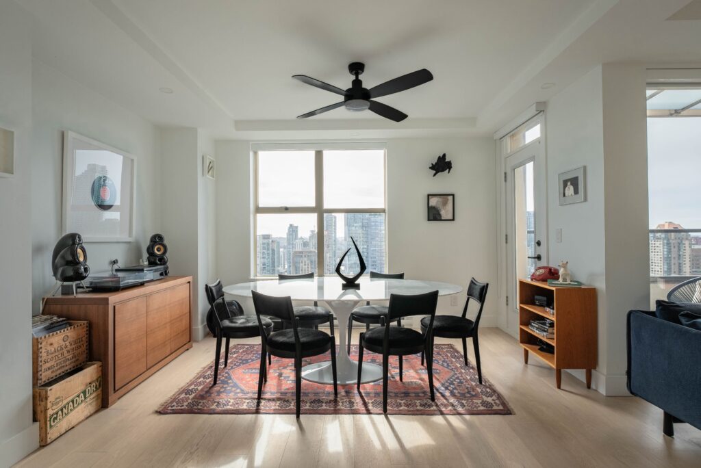 A modern dining room with a round table, six black chairs, large windows showing a city skyline, a ceiling fan, art on white walls, a patterned rug, and wooden cabinets with decor and records.