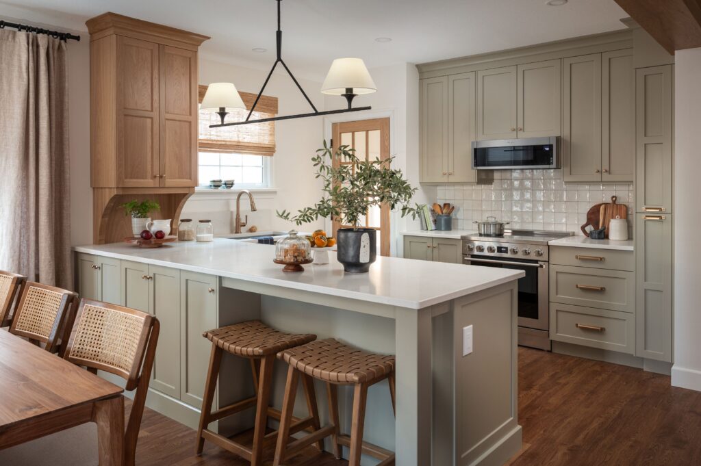 Modern kitchen with light sage green cabinets, a white island with two woven stools, stainless steel appliances, wood accents, a vase with greenery, and a dining area with rattan chairs on a wooden floor.