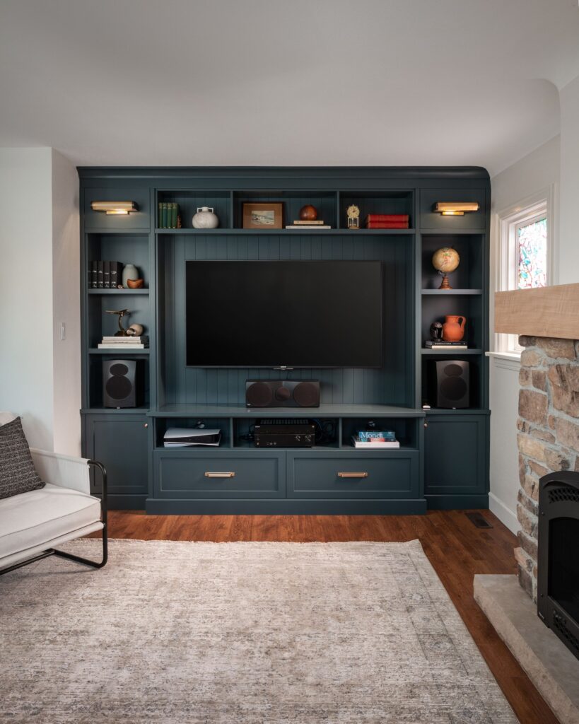 A living room entertainment center with a dark blue built-in unit, a large TV, speakers, books, and decorative items. A stone fireplace is on the right, and a light armchair and rug are on the left.