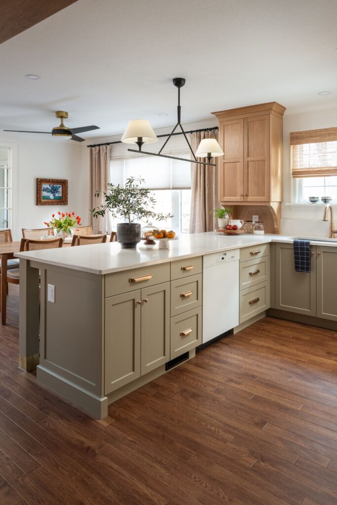 A modern kitchen with sage green cabinets, wooden floors, a large island with gold handles, and two pendant lights. Sunlight streams through windows with light blinds, and plants and fruit decorate the counters.