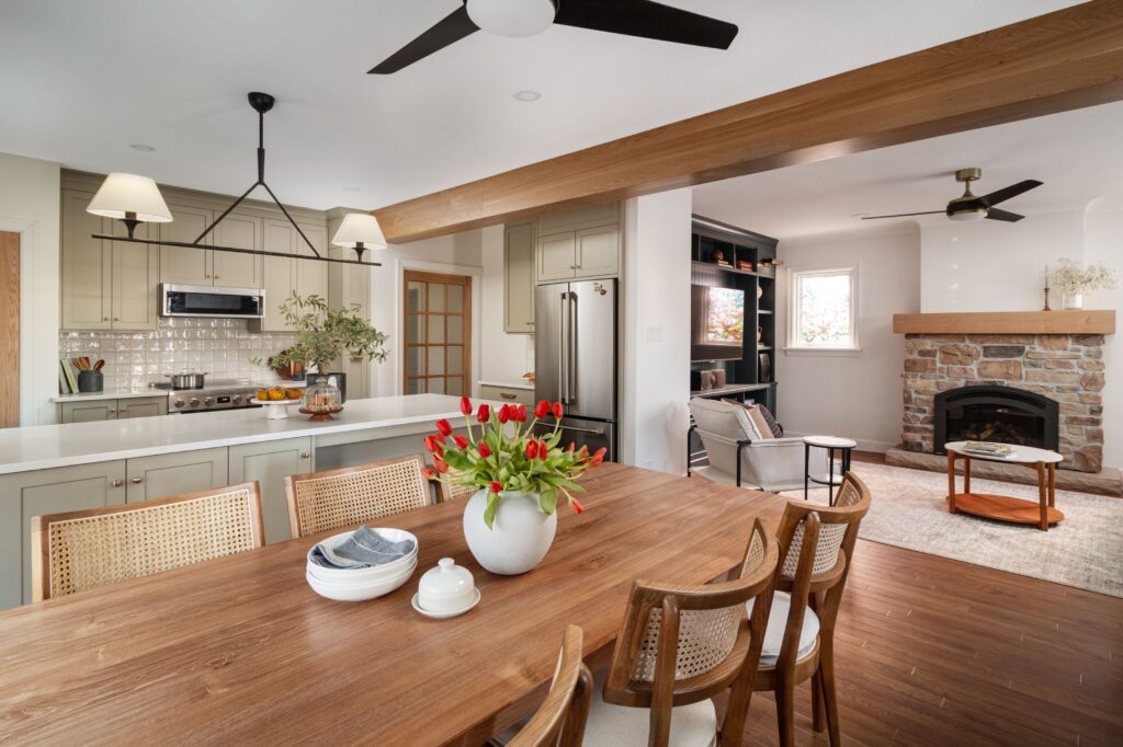 Modern open-concept kitchen and living space with wooden dining table, chairs, a vase of red tulips, light cabinetry, stainless steel appliances, and a cozy stone fireplace in the living area.