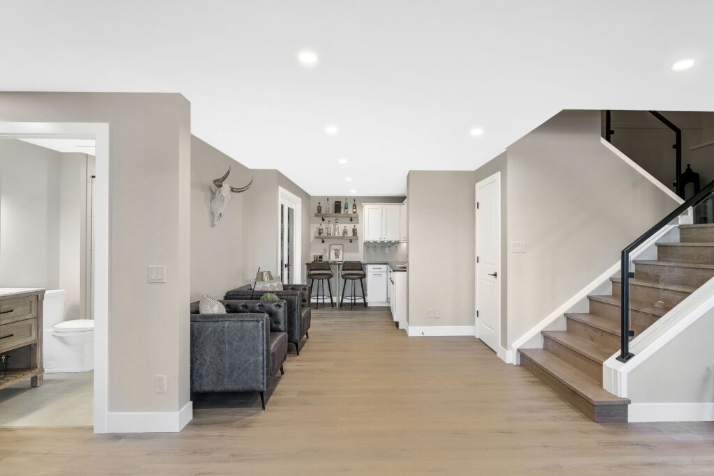 Modern open-concept basement with light wood floors, dark leather armchairs, a mounted antelope head, a small bar with stools, white cabinetry, and a staircase with a glass railing. A bathroom is visible to the left.