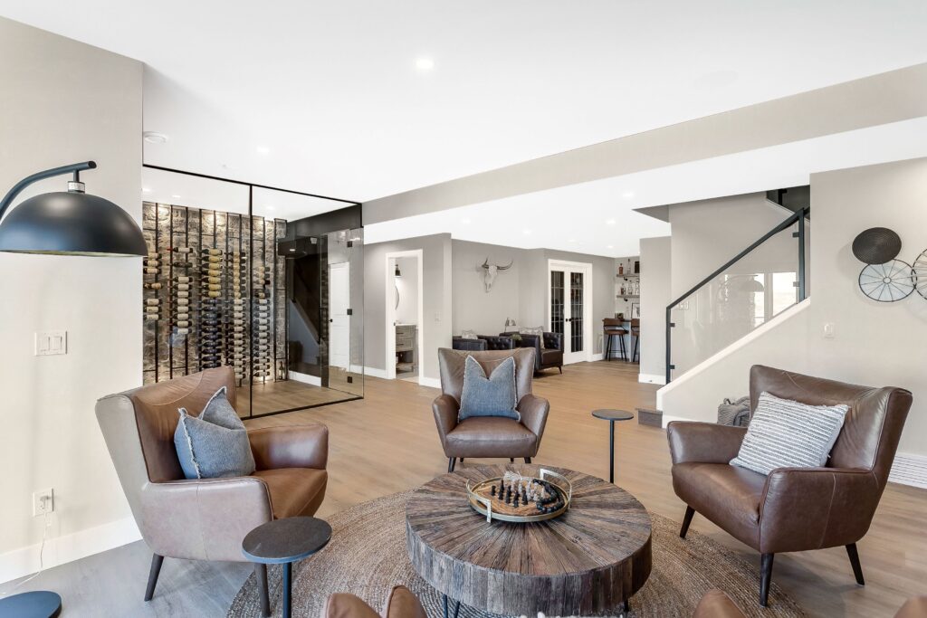 Modern living room with three brown leather armchairs around a wooden coffee table, a chess set on the table, glass-enclosed wine storage in the background, neutral walls, and wooden floors.