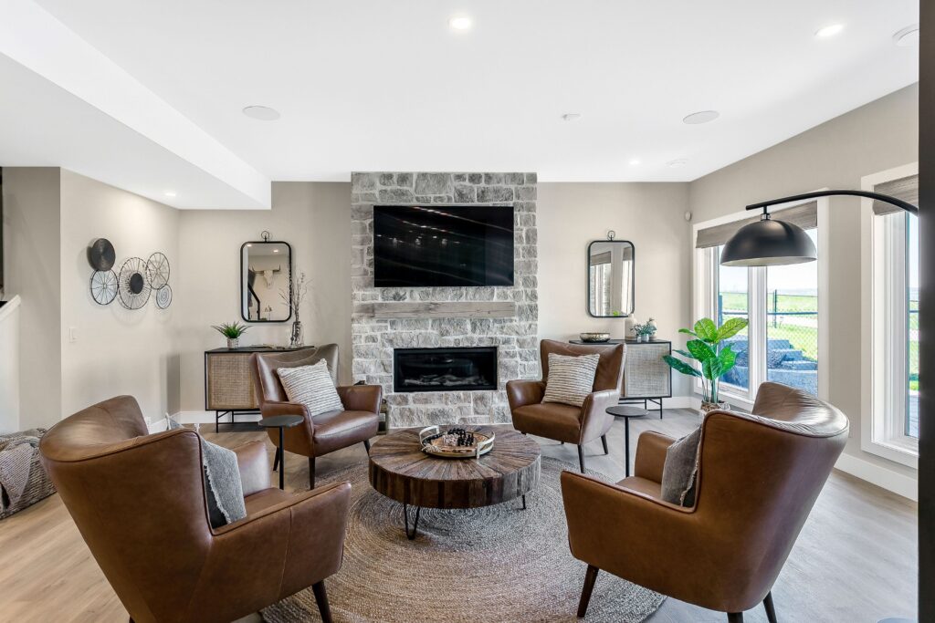 Modern living room with four brown leather chairs arranged in a circle around a round coffee table, a stone fireplace with a mounted TV, large windows, and decorative mirrors on the walls.