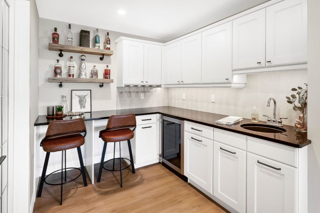 Modern home bar with white cabinets, black countertops, two brown bar stools, open shelves with bottles, a small sink, and decor accents, all set on light wood flooring.