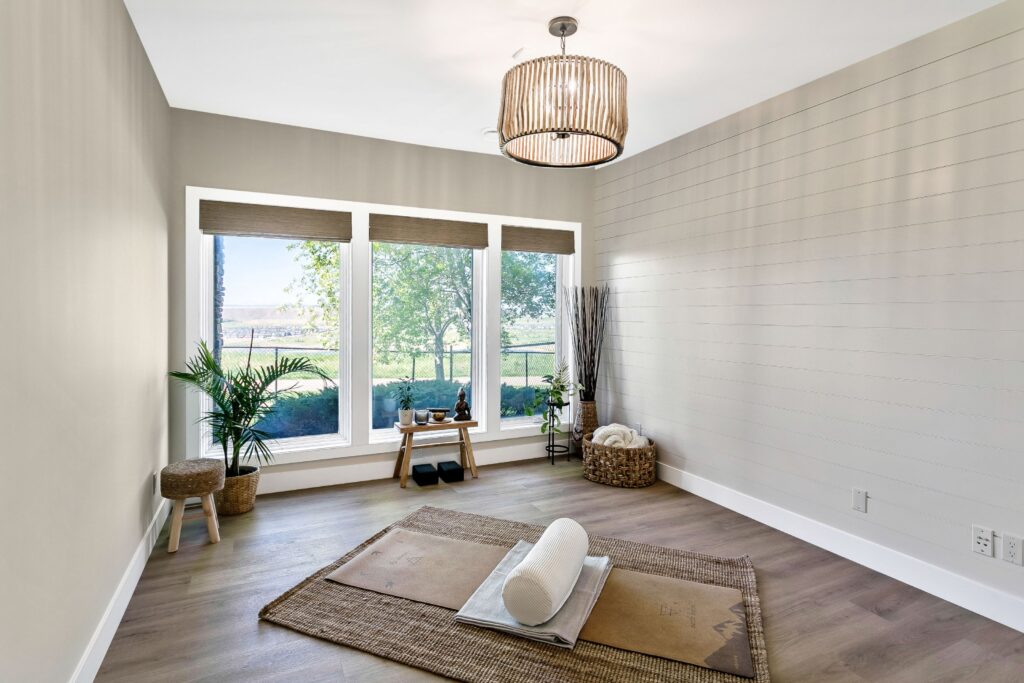 Minimalist yoga or meditation room with large windows, a woven mat, bolster, simple decor, wooden floor, plants, a wicker basket, and calming natural light streaming in.