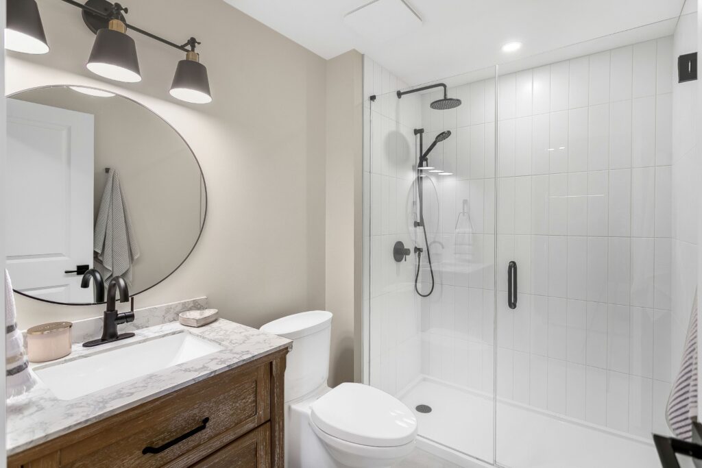 Modern bathroom with a round mirror above a wooden vanity, white sink, and black fixtures. To the right, there is a glass-enclosed shower with white tile walls and a black showerhead. Neutral wall colors and bright lighting.