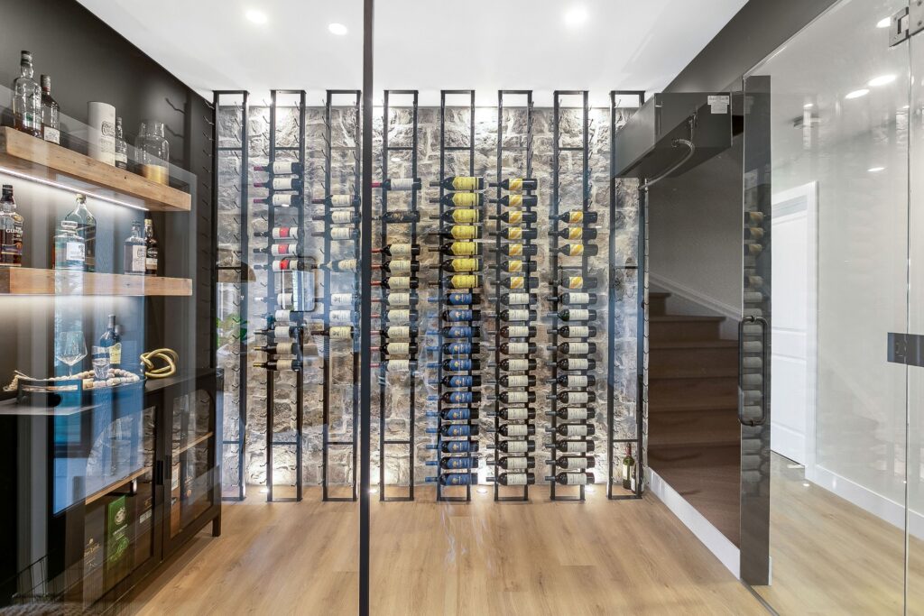 Modern wine cellar with floor-to-ceiling glass walls, wooden floor, and a display of wine bottles on metal racks. Shelves with various liquor bottles are on the left, and a staircase is visible in the background.