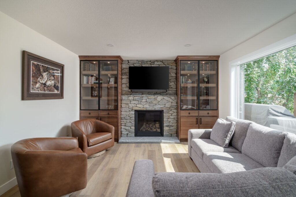 A modern living room with a stone fireplace, wall-mounted TV, glass-front cabinets, two brown leather chairs, a gray sofa, and a large window letting in natural light. A framed animal artwork hangs on the wall.