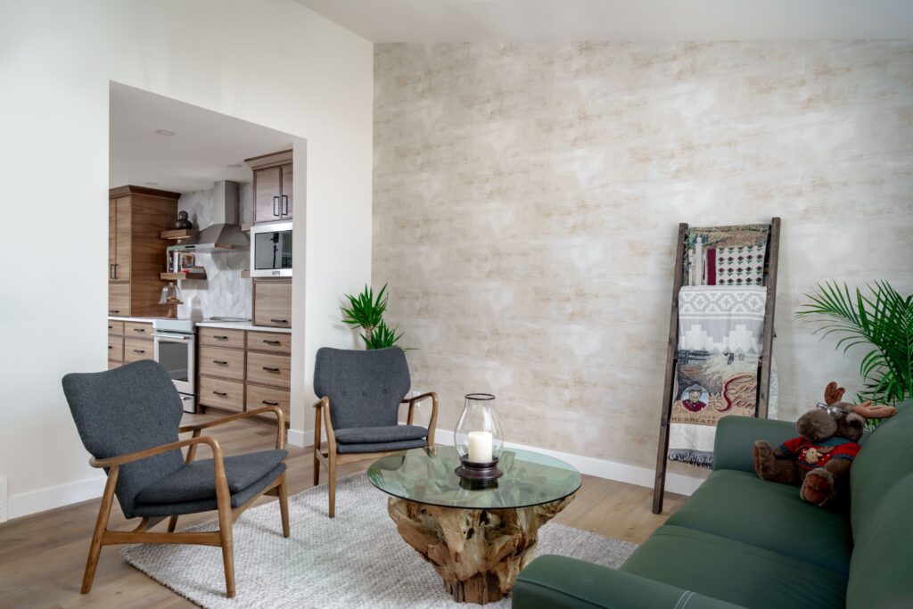Modern living room with two gray armchairs, a green sofa, a glass coffee table on a wooden base, a decorative ladder with blankets, potted plants, and a peek into a wooden kitchen area.