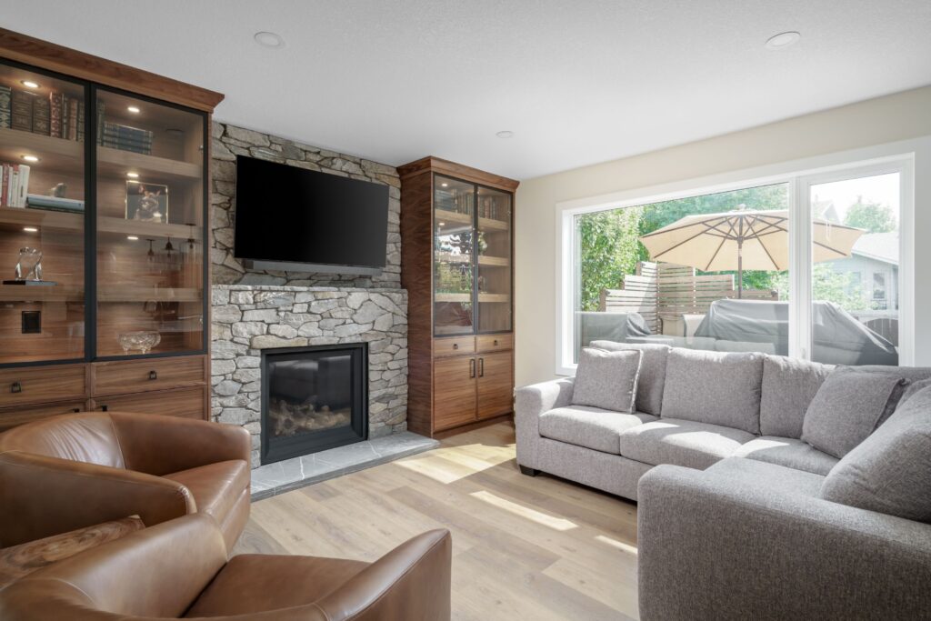 A bright living room with a stone fireplace, wall-mounted TV, wooden built-ins with glass doors, light gray sofa, brown leather chairs, and large window showing a patio with an umbrella outside.