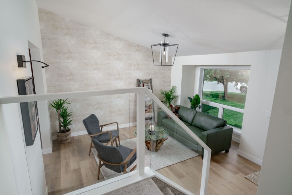 Modern living room with light wood floors, white walls, a green sofa, two gray armchairs, potted plants, and large windows overlooking a grassy yard. A geometric light fixture hangs from the ceiling.