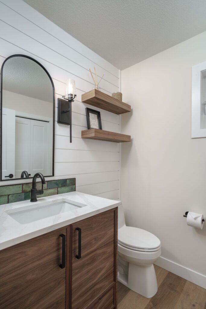Modern bathroom with a wooden vanity, white sink, black faucet, arched mirror, wall sconce, floating shelves with decor, and a toilet with a roll of toilet paper on the wall. White walls and wood floor.