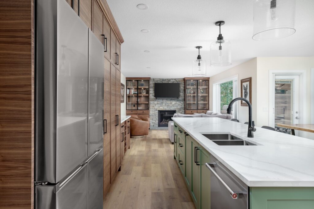 Modern kitchen with stainless steel refrigerator, wood cabinets, green island with sink, marble countertop, pendant lights, and an open view to a living area with a stone fireplace and wall-mounted TV.