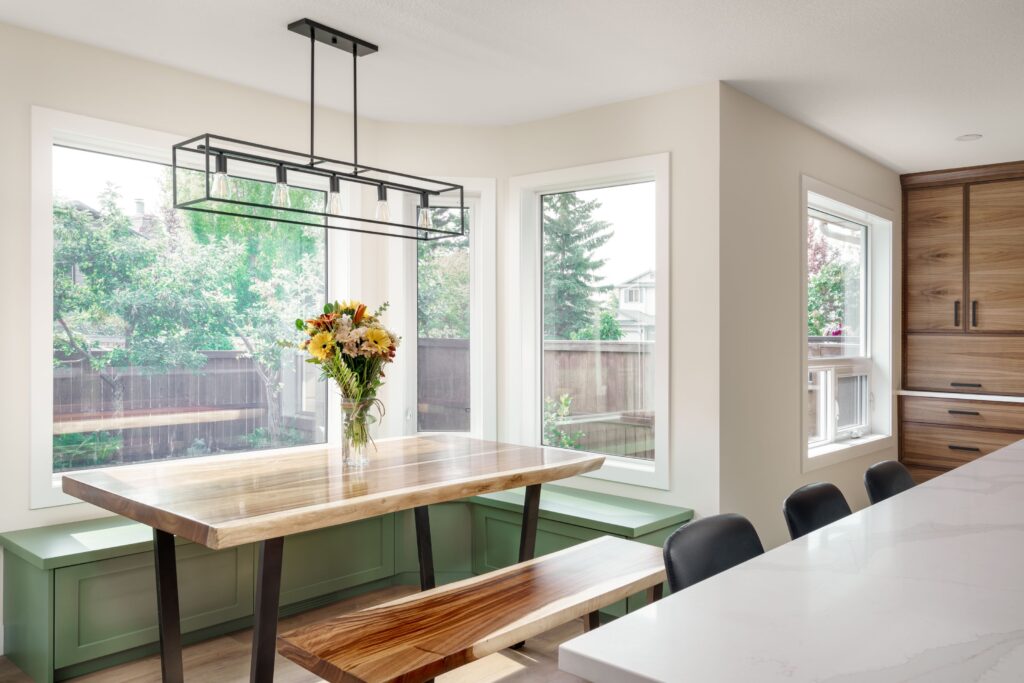A bright dining area with large windows, a wooden table, matching benches, a vase of flowers, green built-in window seating, and modern hanging light fixture. The space has white walls and wood cabinetry.