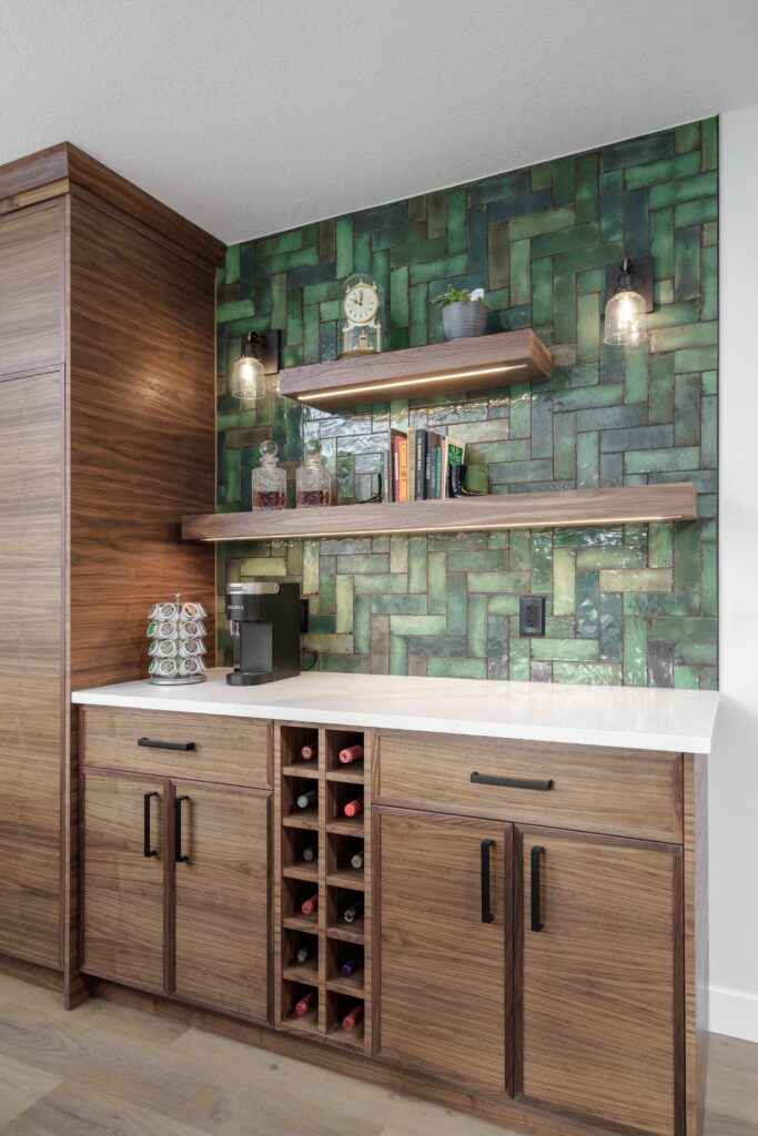 A modern home bar with wooden cabinets, a wine rack, a coffee machine, open shelves with books and decor, and a green herringbone tile backsplash, illuminated by two wall sconces.