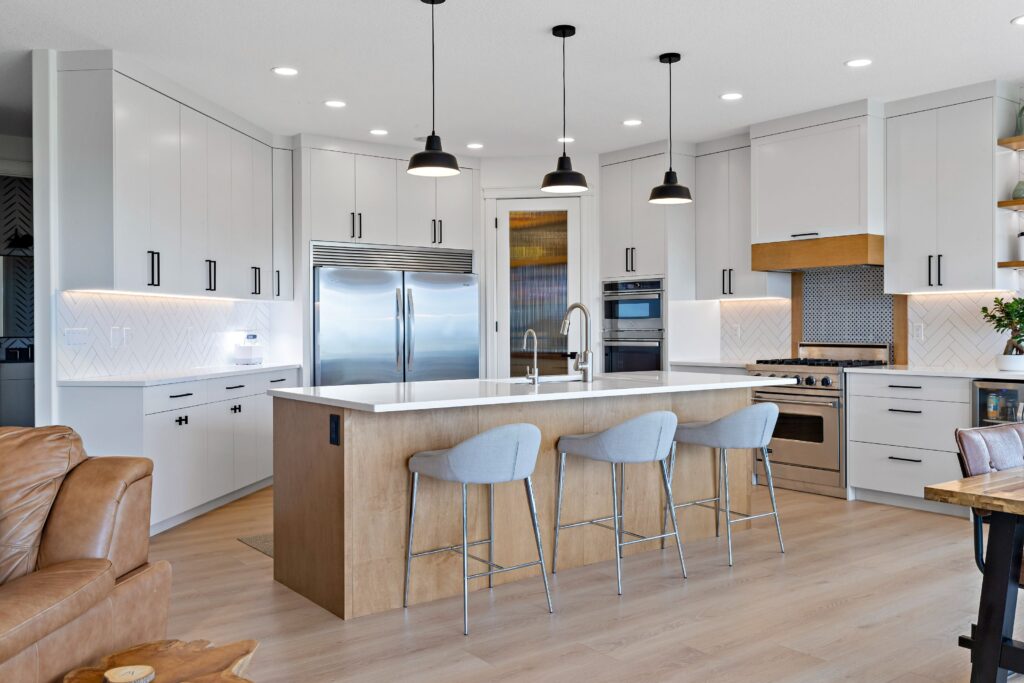 Modern kitchen with white cabinets, stainless steel appliances, a large wooden island with a white countertop, three gray barstools, pendant lights, and light wood flooring.