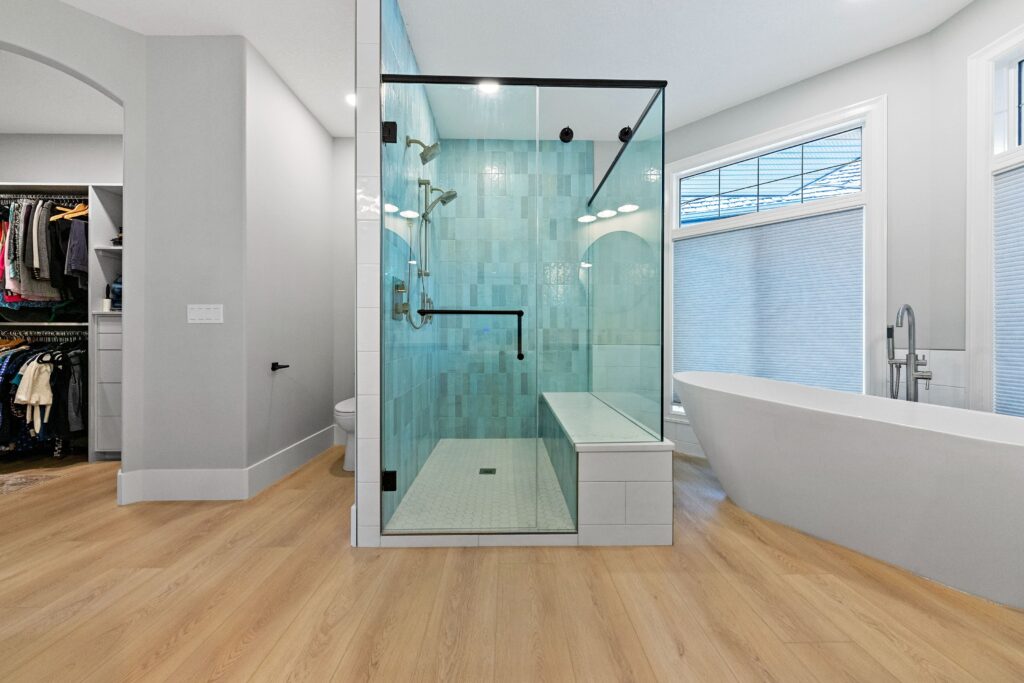 Modern bathroom with light wood floors, a glass-enclosed shower with a bench, a freestanding white bathtub, large windows with blinds, and an open walk-in closet visible in the background.