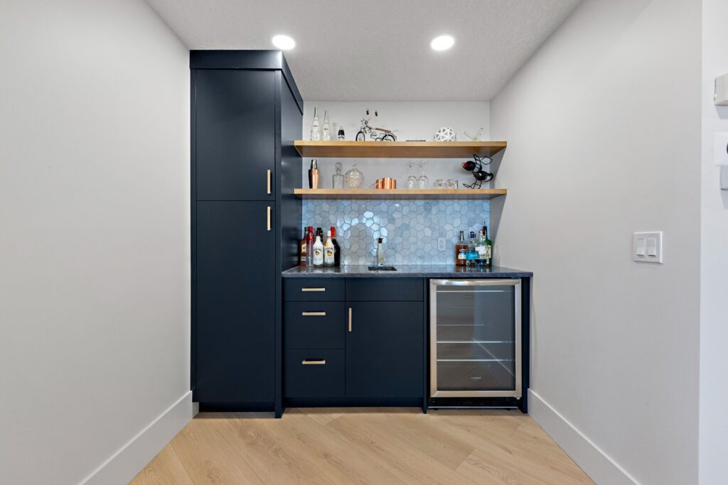 A modern home bar with dark blue cabinets, a small sink, glass shelves holding bottles and glasses, a wine fridge, and a light hexagonal tile backsplash, set against white walls and light wood flooring.