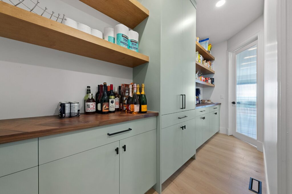 A modern pantry with light green cabinets, open wooden shelves holding paper towels and canned goods, and a countertop with bottles of wine and liquor; a door and more shelves are visible in the background.