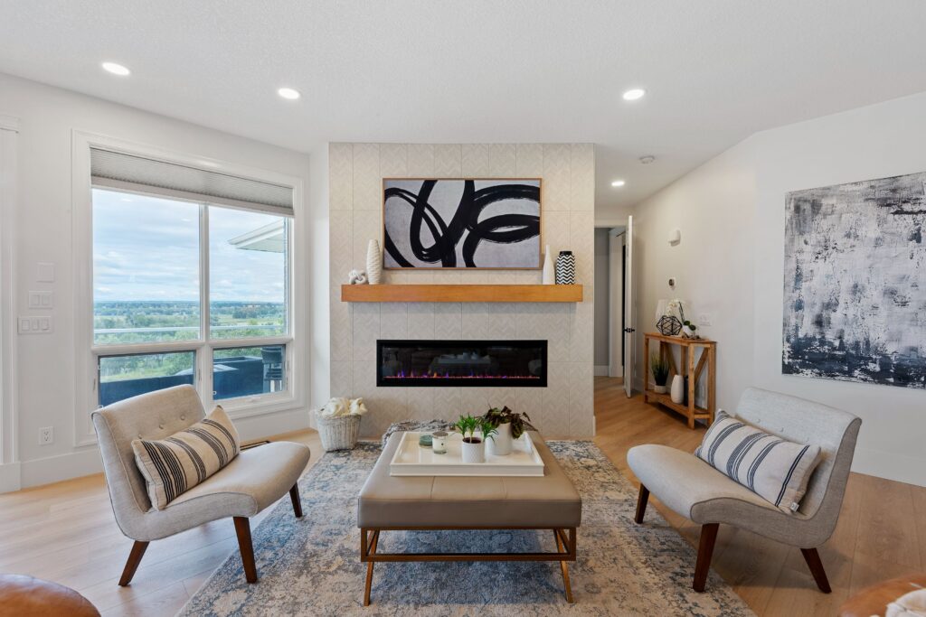 A modern living room with two cushioned chairs, a square ottoman coffee table, plants, and a fireplace set in a patterned wall beneath abstract art. A large window offers a scenic outdoor view.