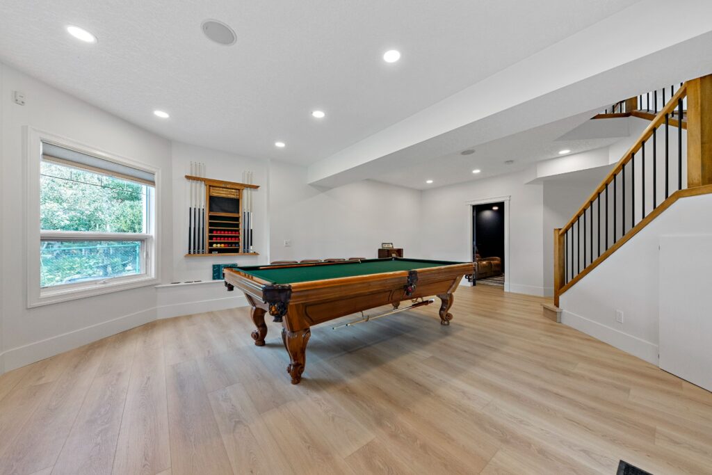 Bright game room with a pool table at the center, cue sticks on the wall, large window with greenery outside, and a staircase with wooden railings leading upstairs. Light wood flooring and white walls enhance the spacious feel.