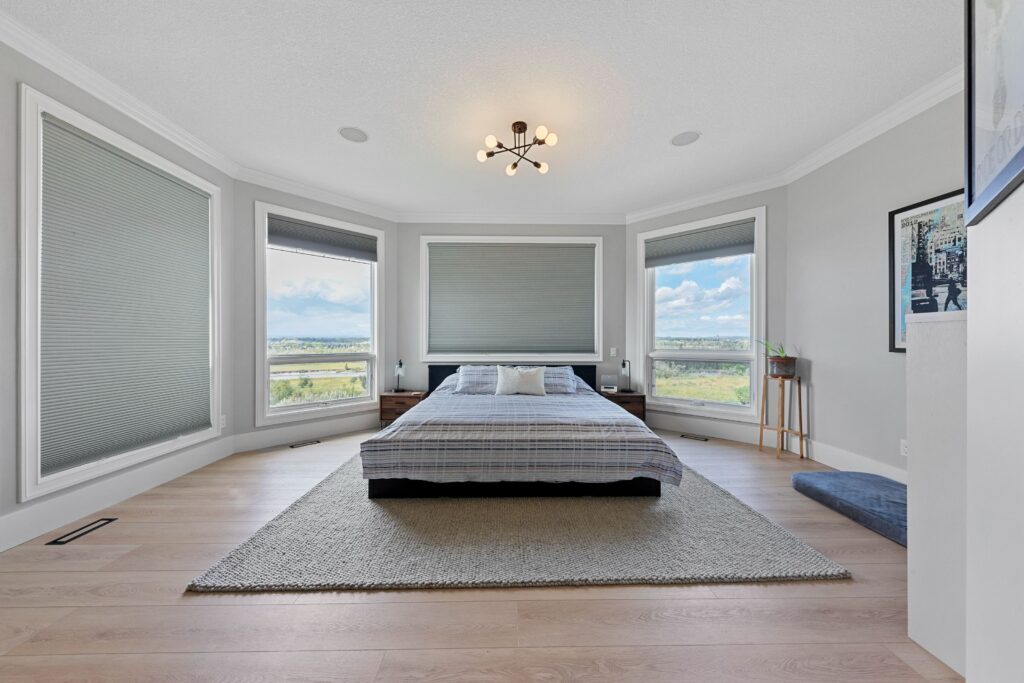 Minimalist bedroom with a low platform bed centered on a rug, large windows with blinds, light wood flooring, and modern ceiling light fixture. The room has a bright, airy feel and a scenic outdoor view.