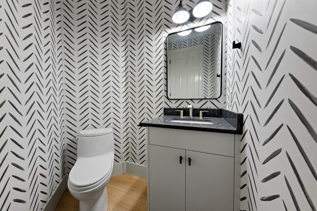 A modern bathroom with bold black-and-white chevron-patterned wallpaper, a white toilet, a vanity with a black countertop and cabinet, a rectangular mirror, and two round lights above the mirror.