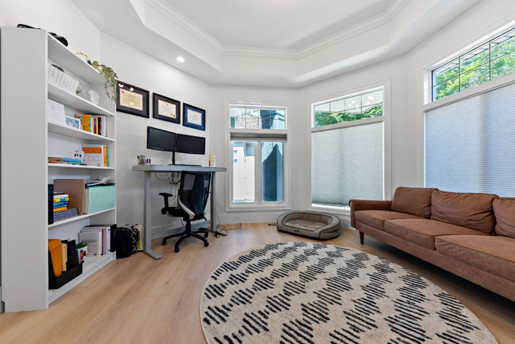 A bright, modern home office with a brown couch, a round patterned rug, a standing desk with dual monitors, a white bookshelf, large windows, and a dog bed by the windows.