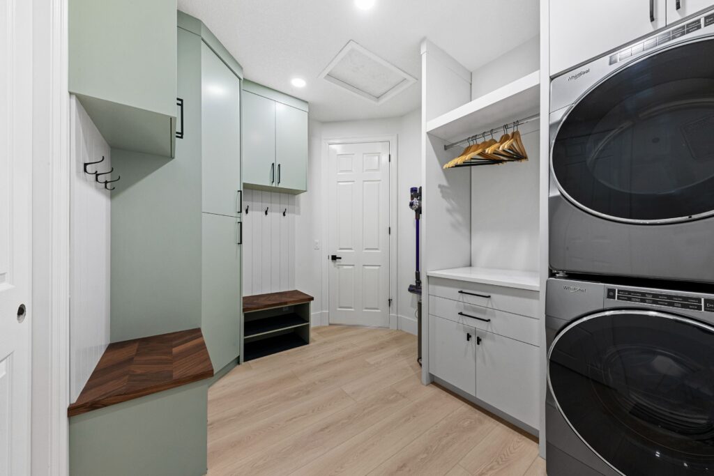 Modern laundry room with light wood floors, stacked washer and dryer, mint green cabinets, wooden benches, hooks, open shelving with hangers, and a white door in the center background.
