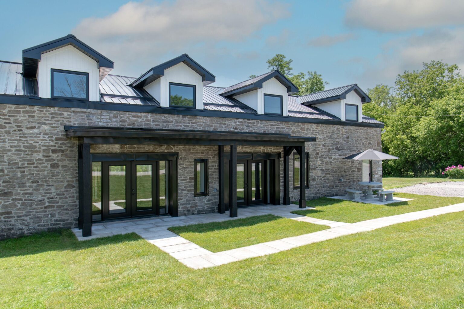 A stone building with a dark metal roof and four dormer windows, large glass doors, a stone patio, and a picnic table with an umbrella on a grassy lawn under a partly cloudy sky.