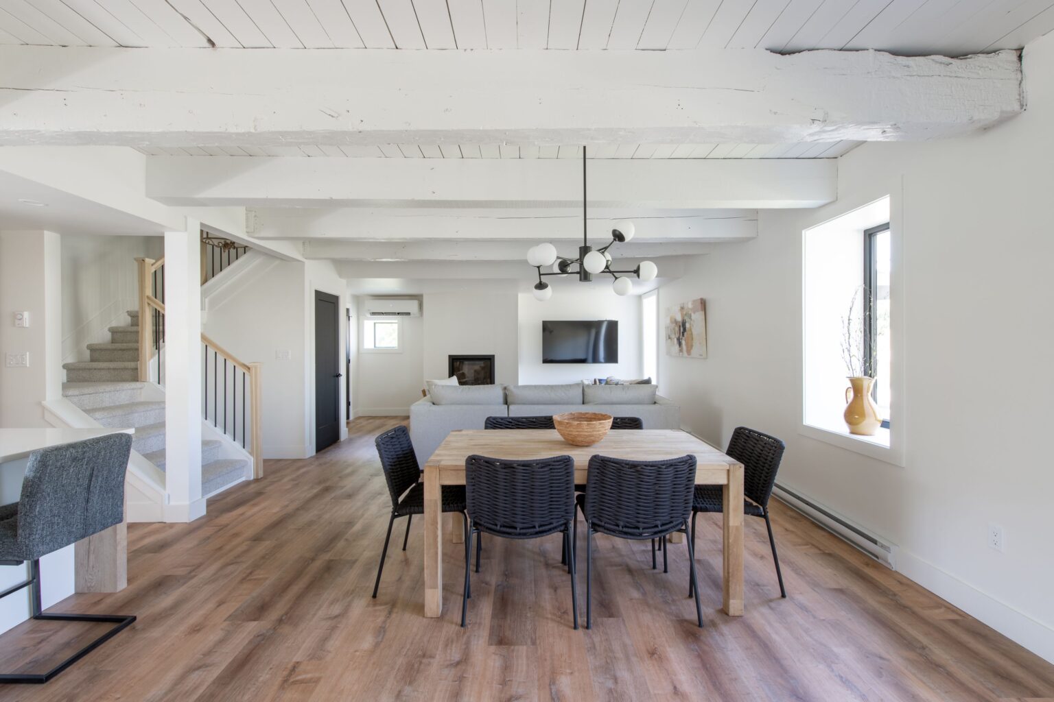 A modern dining area with a wooden table, six black woven chairs, a statement chandelier, wood floors, white walls, exposed ceiling beams, and large windows letting in natural light. A living area and staircase are visible in the background.
