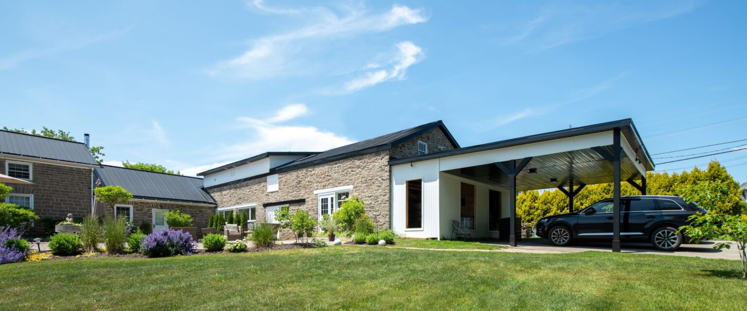 An older house turned modern with stone and white walls, black roof, and attached carport with a black SUV parked underneath. The house is surrounded by a green lawn, shrubs, and purple flowers under a blue sky.