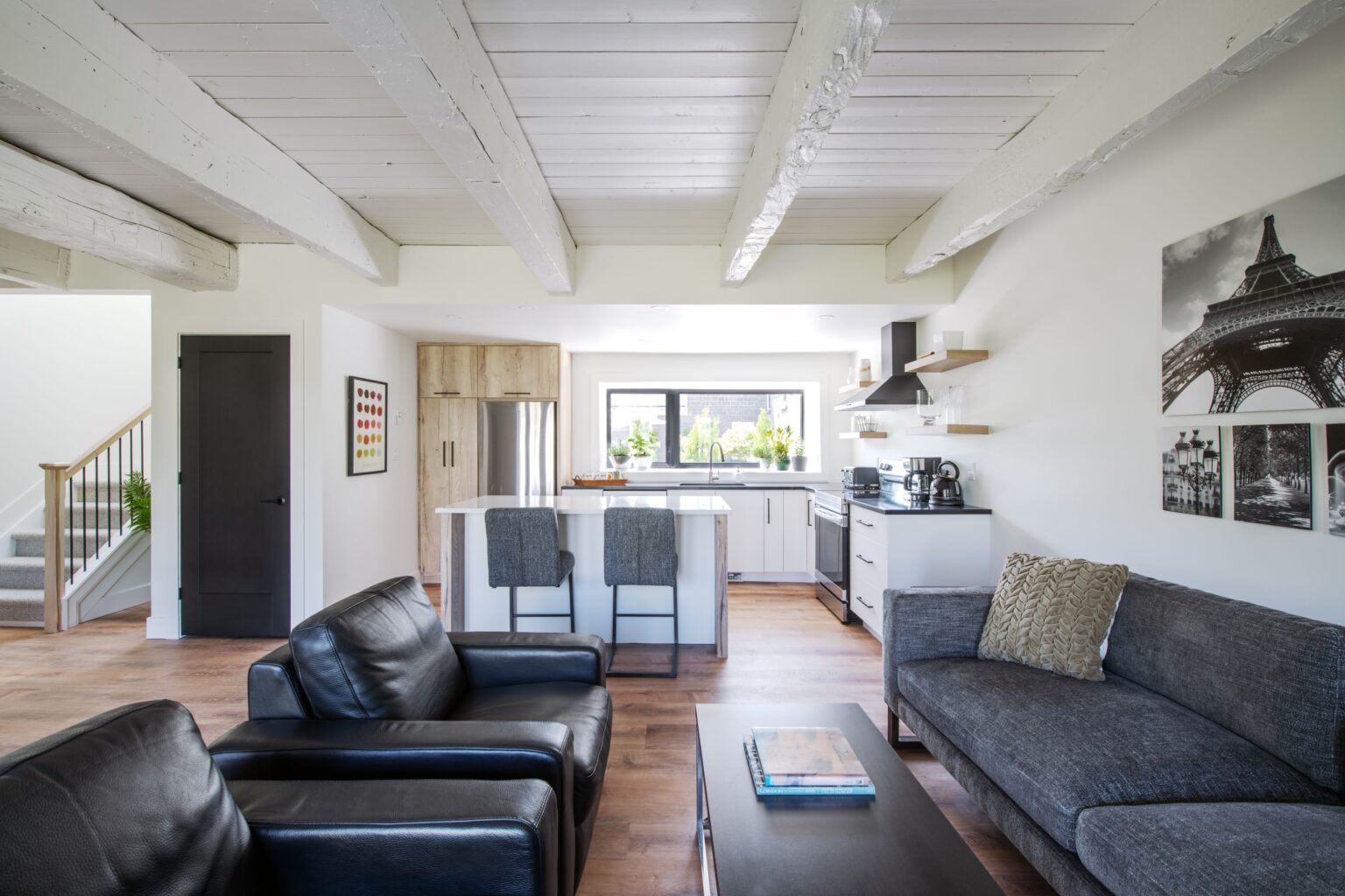 Modern open-plan living room and kitchen with white walls, exposed ceiling beams, black leather chairs, a gray sofa, wood floors, and a kitchen island with stools. Eiffel Tower art is displayed on the wall.