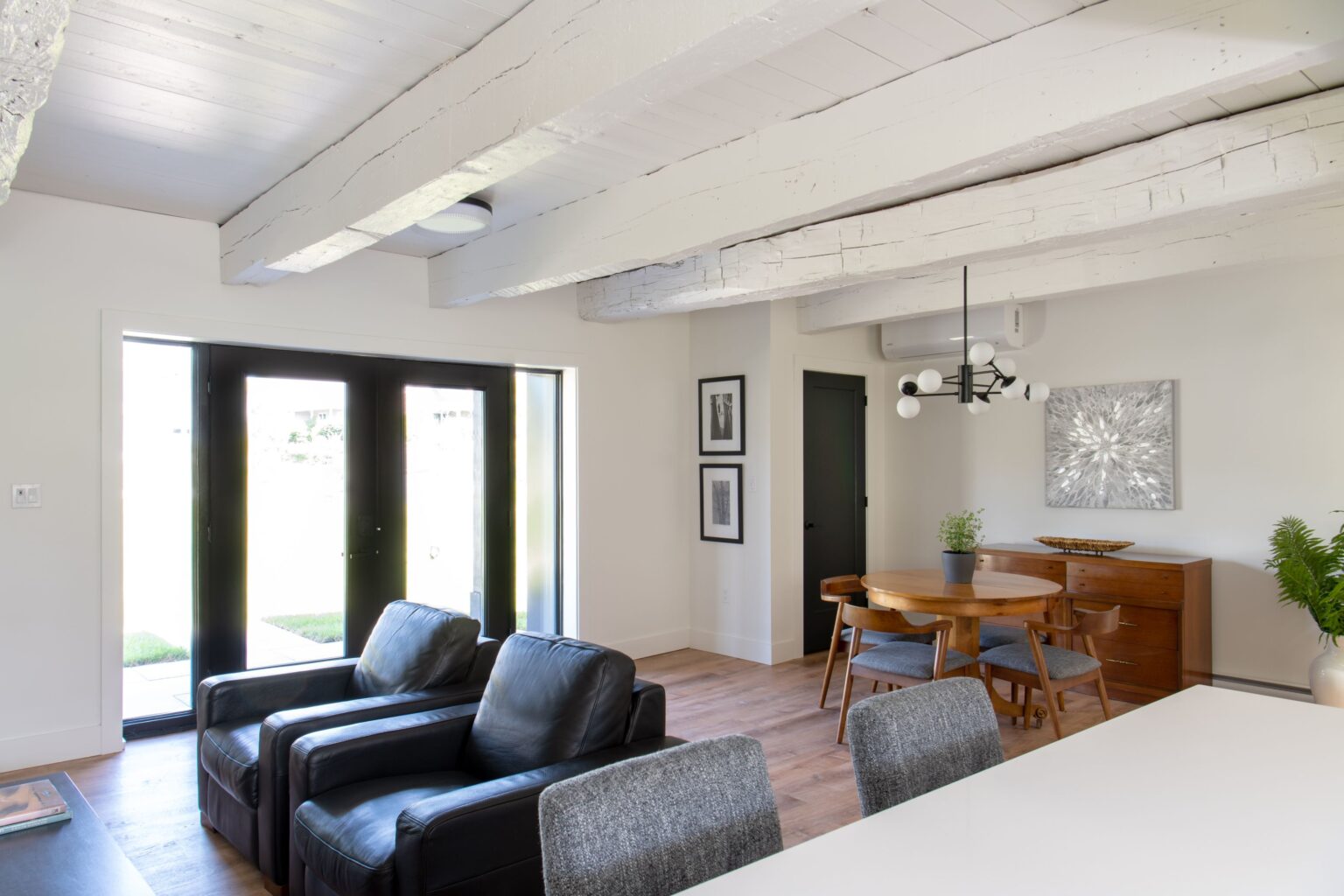 A bright, modern living and dining area features black leather chairs, a round wooden dining table, mid-century modern furniture, exposed white ceiling beams, and large double glass doors letting in natural light.