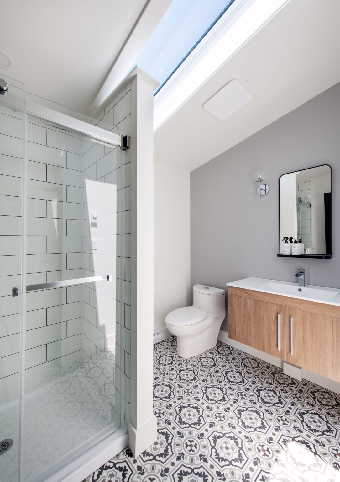 Modern bathroom with patterned tile floor, glass-enclosed shower with white subway tiles, light wood vanity with sink and mirror, toilet, and skylight letting in natural light.