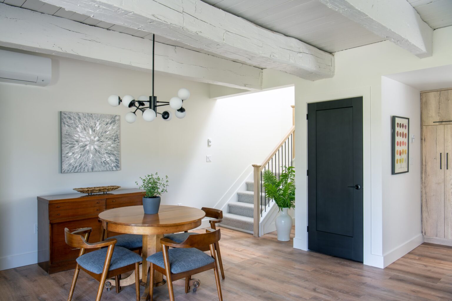 A modern dining area with a round wooden table, four chairs, a potted plant, and a contemporary chandelier. In the background are stairs, a black door, a wooden sideboard, artwork, and a large potted fern.