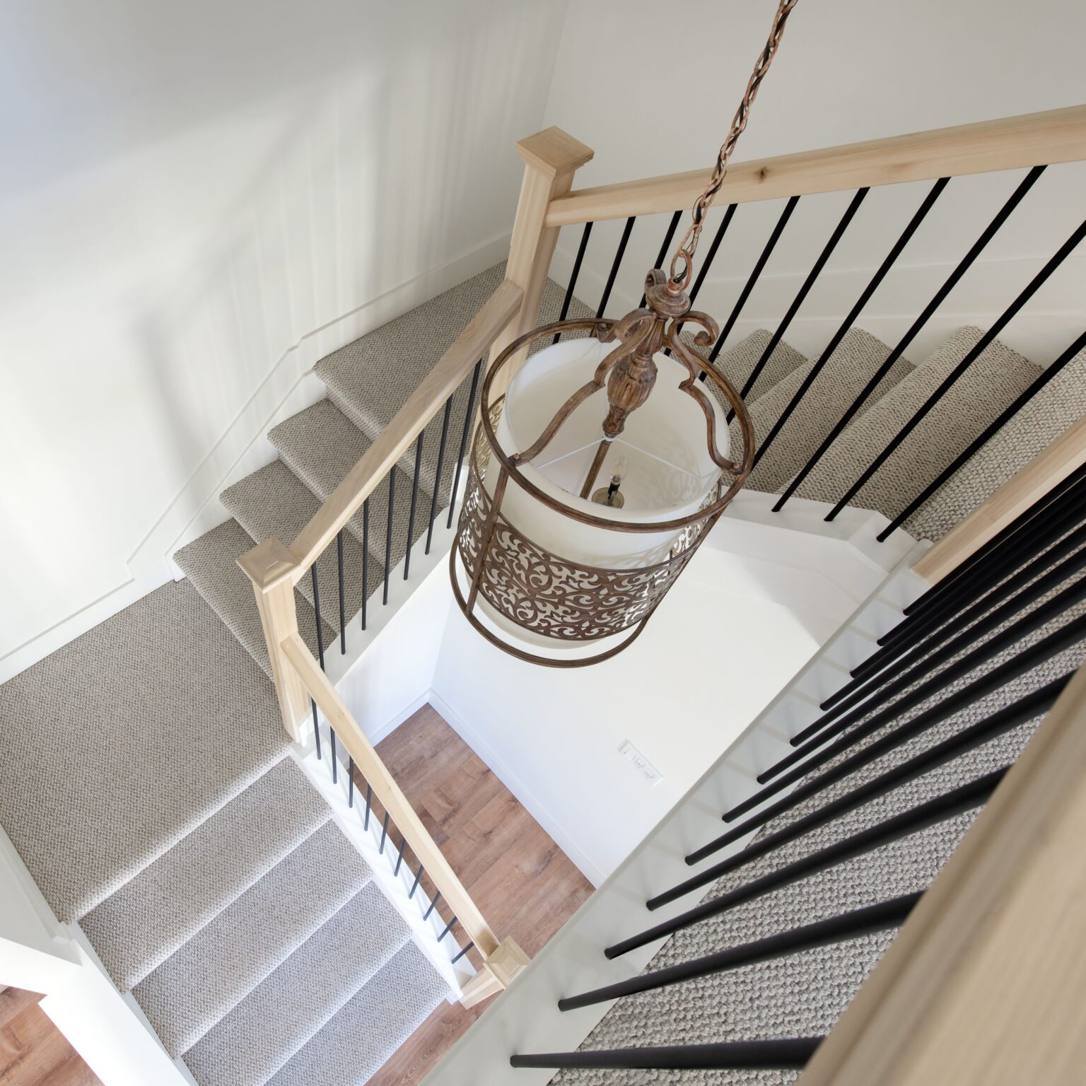 View from above of a carpeted staircase with light wood handrails and black balusters, featuring a decorative hanging lantern light fixture. The stairs turn at a landing with wood flooring below.