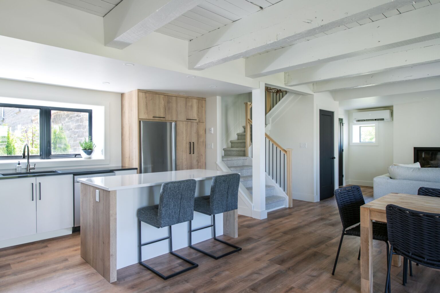 Modern kitchen and living space with white walls, wooden floors, exposed ceiling beams, an island with two gray stools, stainless steel fridge, dining table, and a staircase leading upstairs. Natural light fills the room.