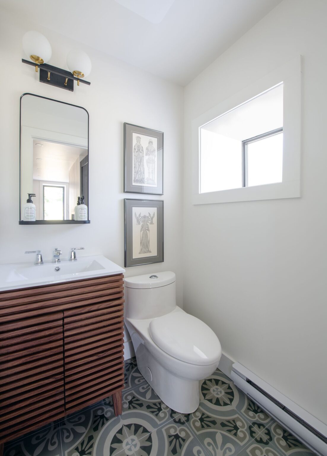 A modern bathroom with a wood slat vanity, rectangular mirror, white sink, and toilet. Two black-and-white framed art prints hang on the wall above the toilet. Patterned tile flooring and a small window provide natural light.