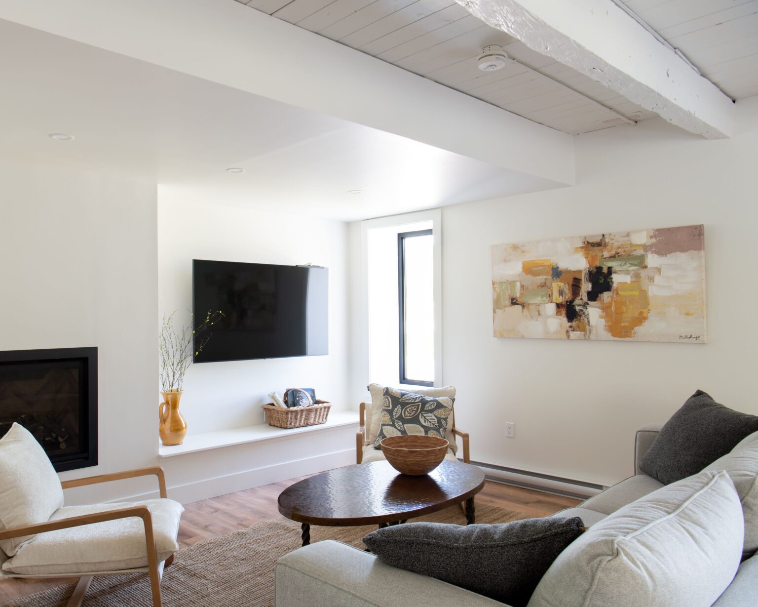 A bright, modern living room with white walls, a mounted TV, abstract artwork, beige armchairs, a gray sofa, a wooden coffee table, and a fireplace. Natural light enters through a tall window.