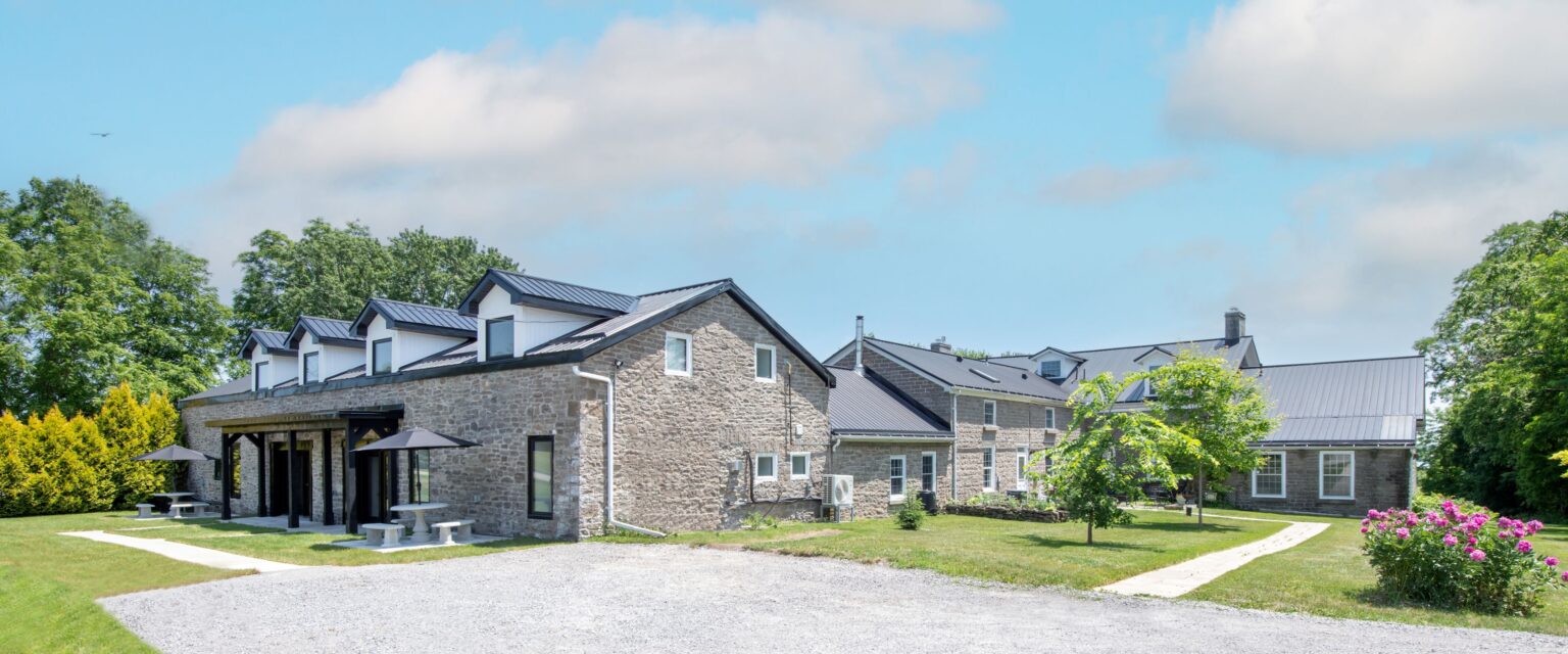 A large stone building with black metal roofs sits on a grassy lawn, surrounded by trees and flowering bushes. There are outdoor tables with umbrellas and a gravel driveway in front under a partly cloudy sky.