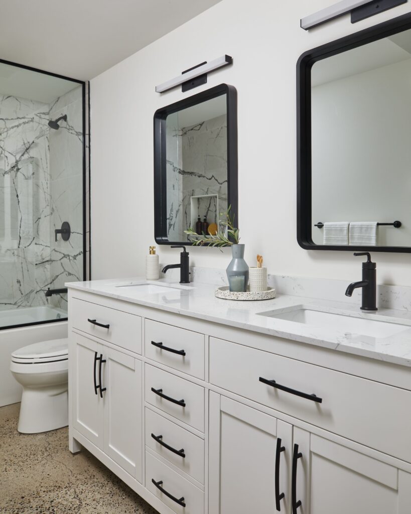 Modern bathroom with a white double-sink vanity, black fixtures, two rectangular mirrors, and a marble shower with glass doors. Decor items and towels are neatly arranged on the counter.