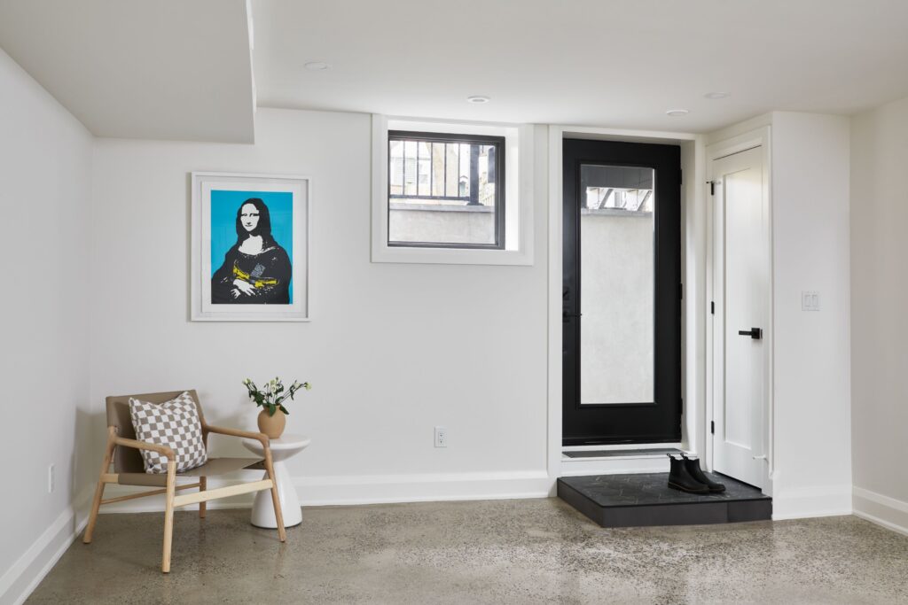 A modern, minimal entryway with a black front door, a small round table with a vase and flowers, a wooden chair with a checkered pillow, and a colorful pop art Mona Lisa print on the white wall.