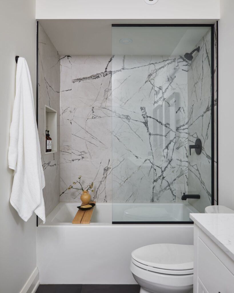 A modern bathroom with a white bathtub and glass shower door, featuring marble-patterned tiles, a black shower fixture, a wooden bath tray with a vase, a towel hanging on the wall, and a white toilet and vanity.