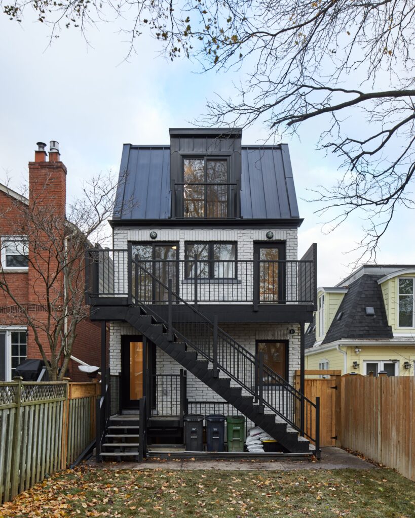 A modern three-story house with white brick exterior, black metal roof, and an outdoor black metal staircase, seen from the backyard. The yard is fenced with some trees and neighboring houses visible.