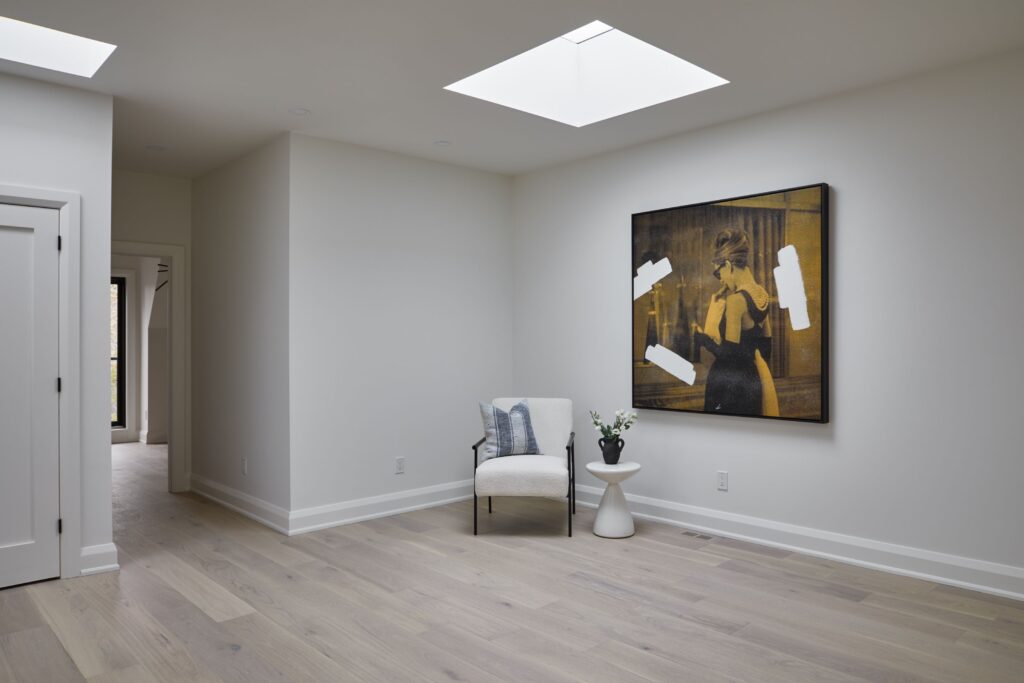 Minimalist room with light wood floors, white walls, a skylight, a modern armchair with a pillow, a small side table with a plant, and a contemporary portrait painting on the wall.