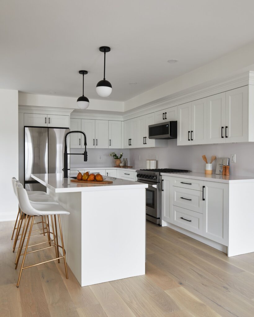 Modern white kitchen with a central island, three white barstools with gold legs, stainless steel appliances, black fixtures, light wood flooring, and two round pendant lights hanging over the island.