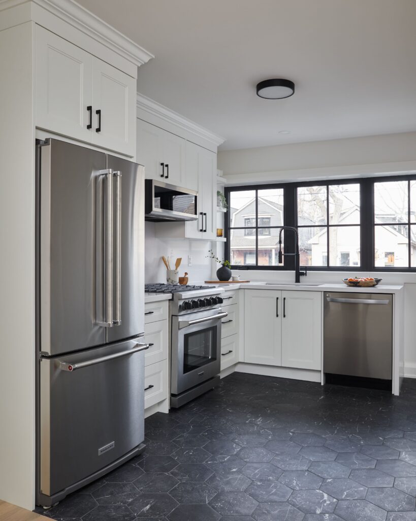 Modern kitchen with white cabinets, stainless steel appliances, black hexagon tile floor, large windows above the sink, and minimalistic decor. Light fills the space from the window.